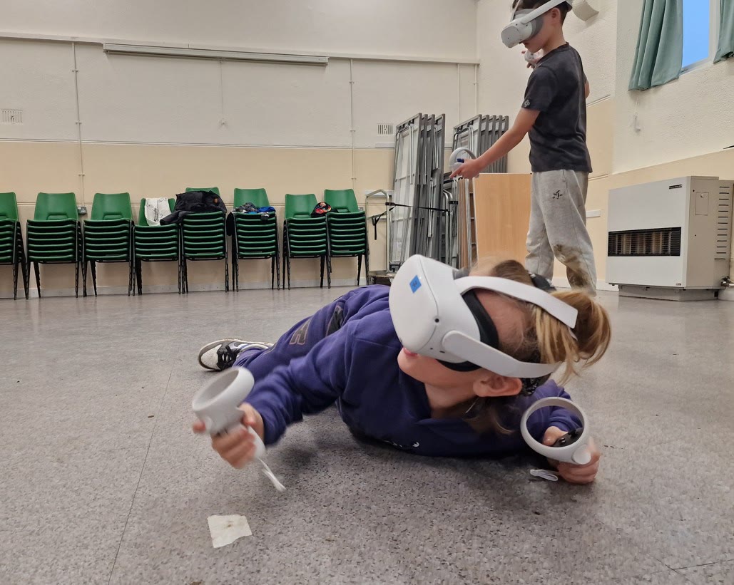 A girl in a VR headset dives flat on a community hall floor mid-game while a boy plays behind her